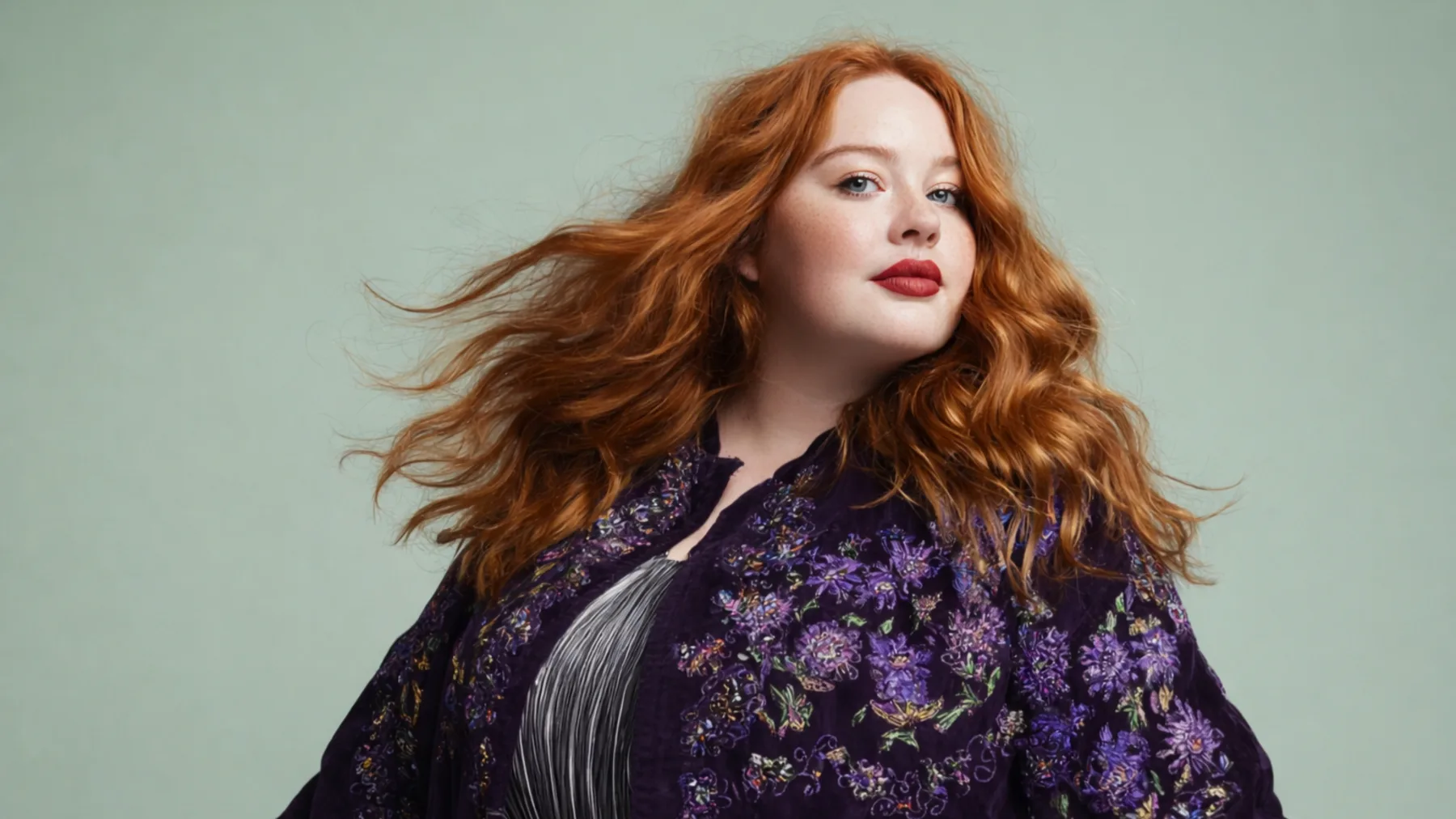 A woman with long auburn hair wearing a richly embroidered deep purple jacket, photographed against a sage green background