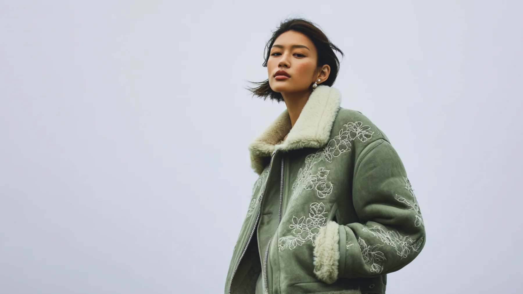 A woman wearing an oversized sage green shearling jacket with floral embroidery, facing forward against a soft grey background