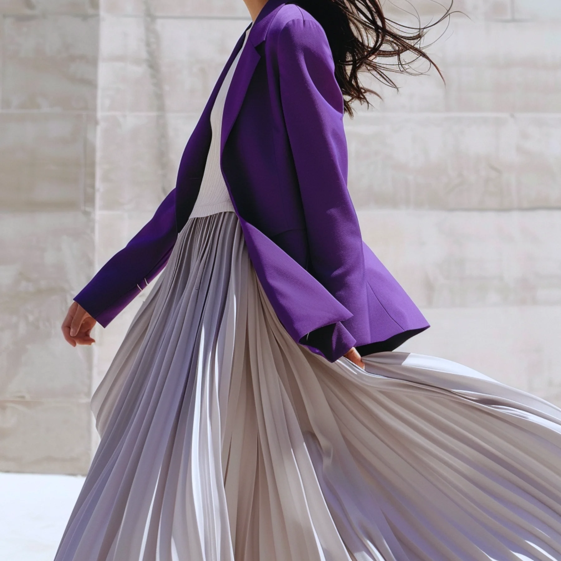 Woman in a deep purple blazer and platinum pleated skirt mid-stride — the feeling of dressing with intention and moving through the world with confidence