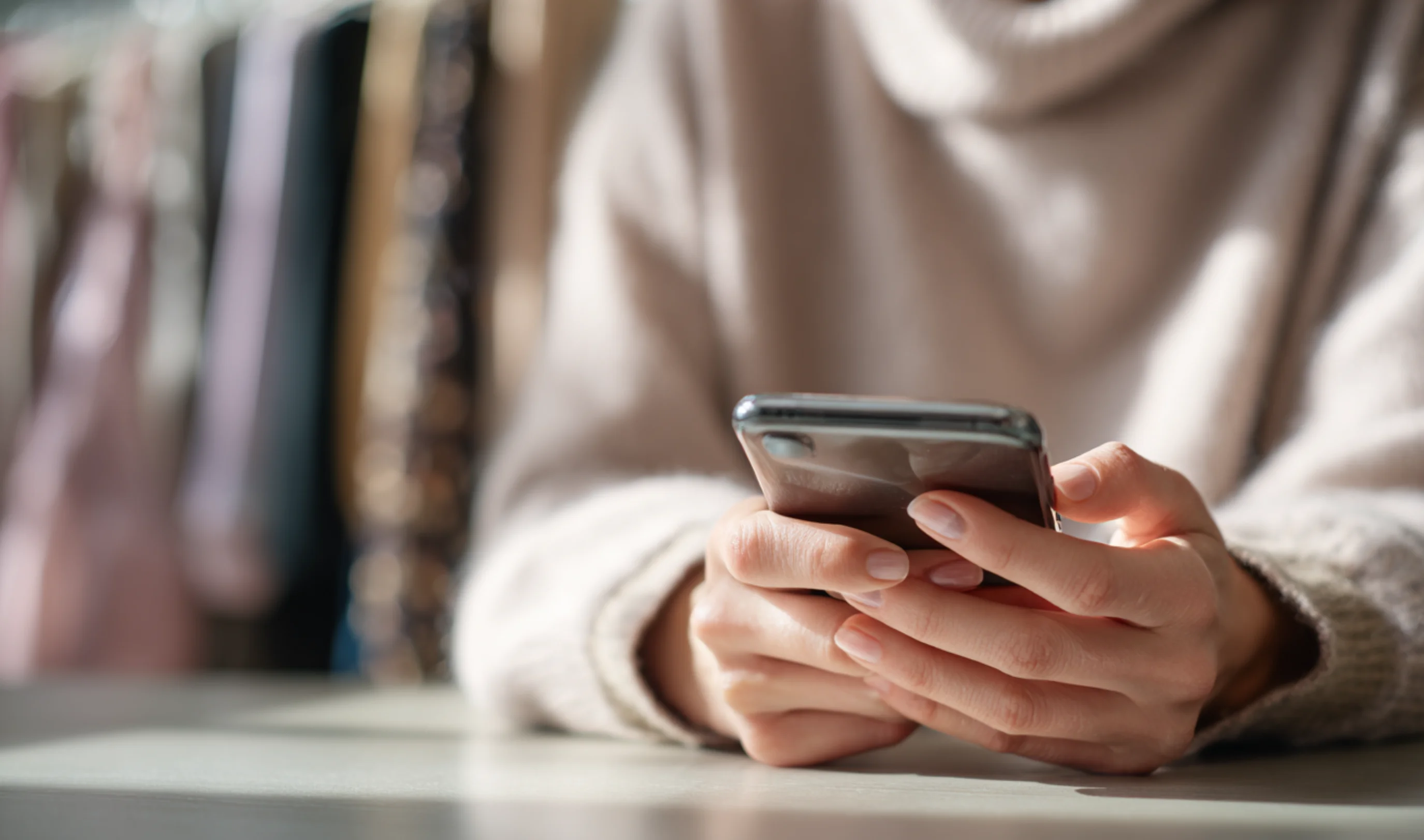 Hands holding a smartphone on a table, with blurred clothing rail in the background